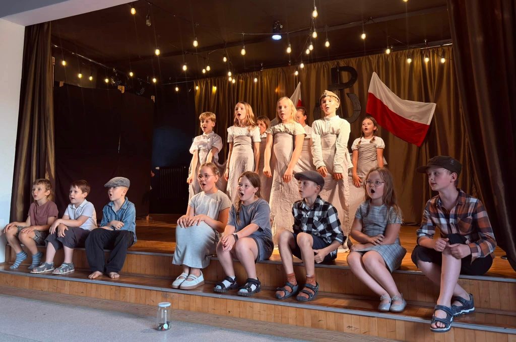 Grupa dzieci występuje na scenie, prezentując przedstawienie o tematyce patriotycznej przed zgromadzoną publicznością.