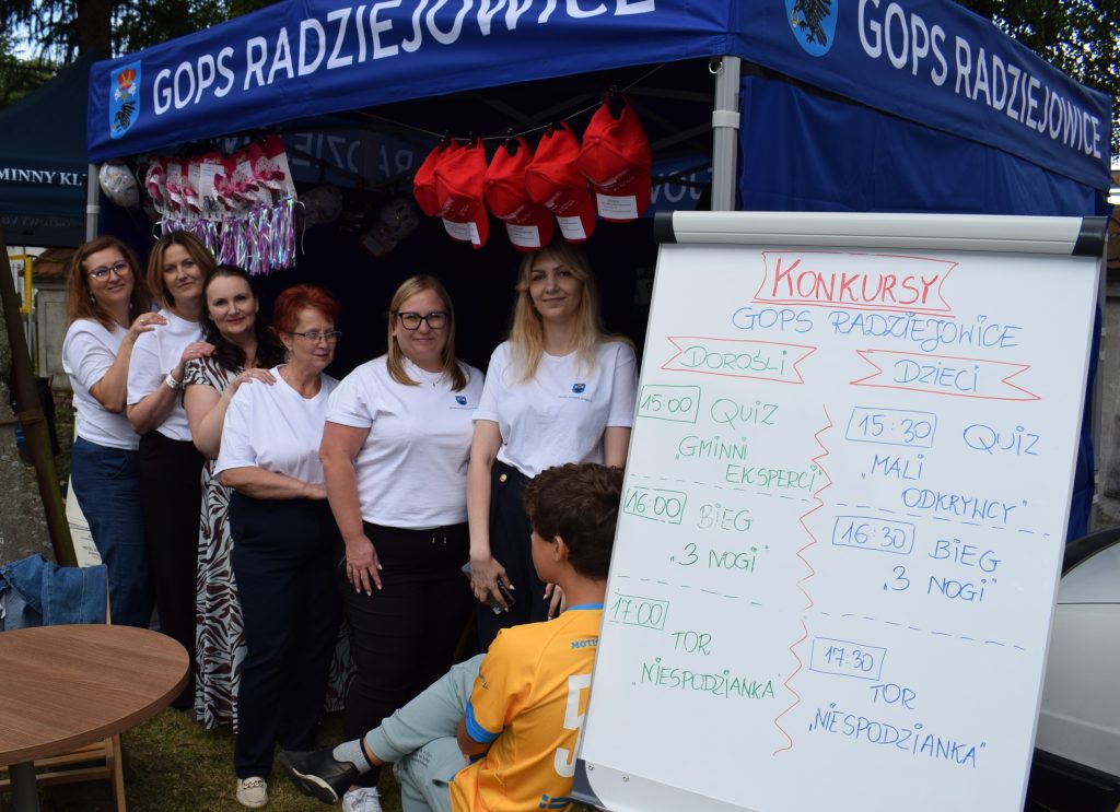 Pracownicy Gminnego Ośrodka Pomocy Społecznej w Radziejowicach stoją przed swoim stoiskiem podczas Święta Chleba. Obok znajduje się tablica z programem konkursów dla dzieci i dorosłych.