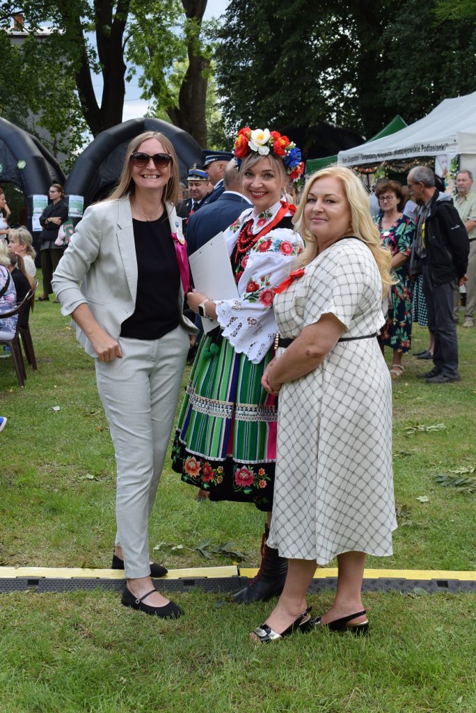 Trzy kobiety pozują do zdjęcia podczas Święta Chleba. Jedna z nich ma na sobie tradycyjny strój ludowy z kolorowym wiankiem, a w tle widoczni są uczestnicy wydarzenia i stoiska.