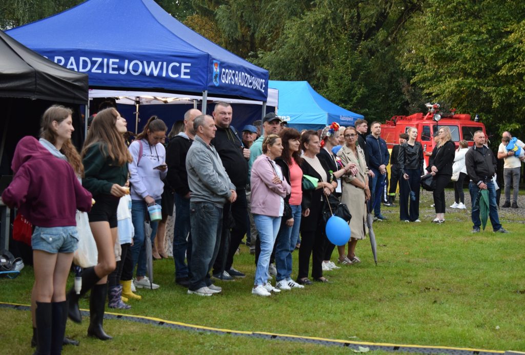 Grupa uczestników stoi przed stoiskiem Gminnego Ośrodka Pomocy Społecznej w Radziejowicach i obserwuje wydarzenia odbywające się na terenie Święta Chleba. W tle widoczny jest namiot oraz wóz strażacki.