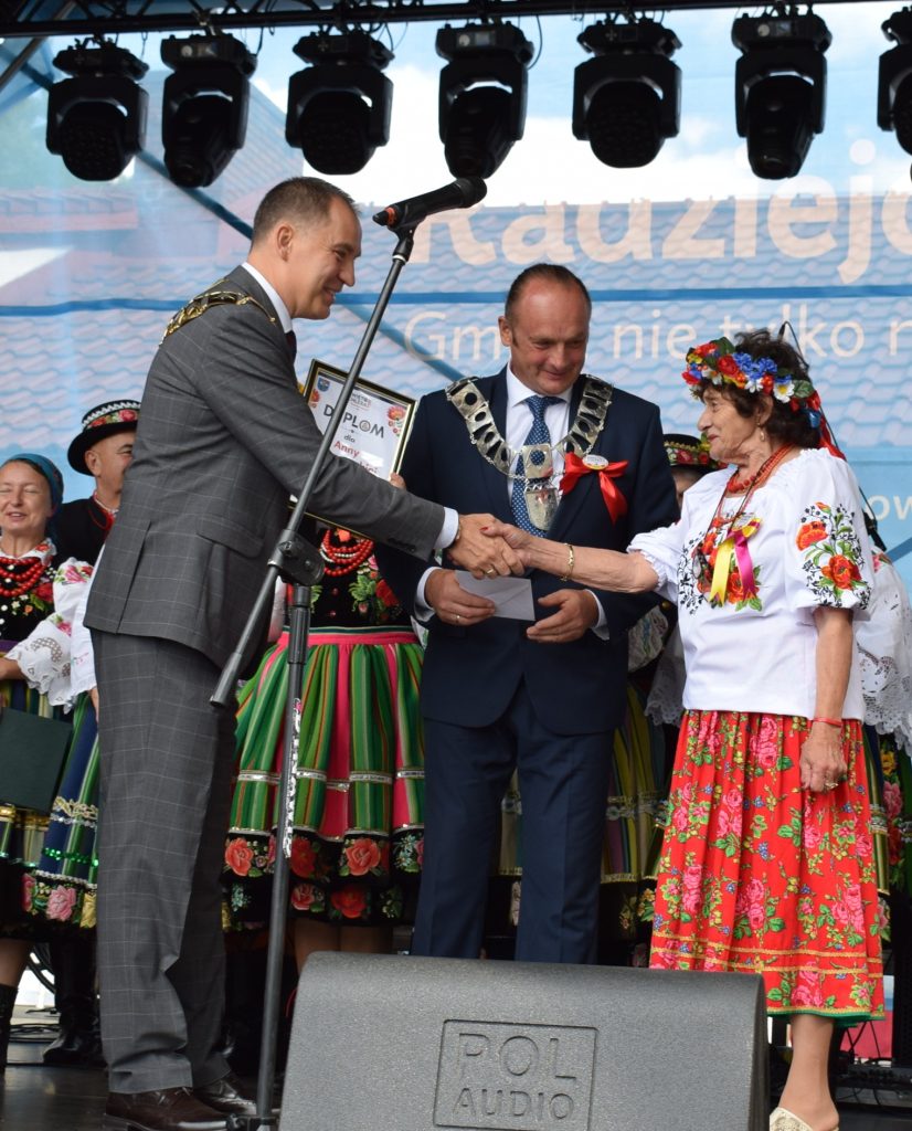 Przedstawiciele organizatorów i uczestnicy uroczystości stoją na scenie podczas wręczania dyplomów i podziękowań. W tle widoczna jest scena z oświetleniem i banerem Radziejowice.