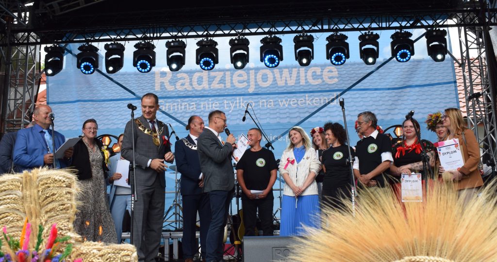 Uczestnicy Święta Chleba spacerują między stoiskami gastronomicznymi i promocyjnymi. Na trawniku bawią się dzieci, a w tle widoczne są liczne namioty i stoiska.