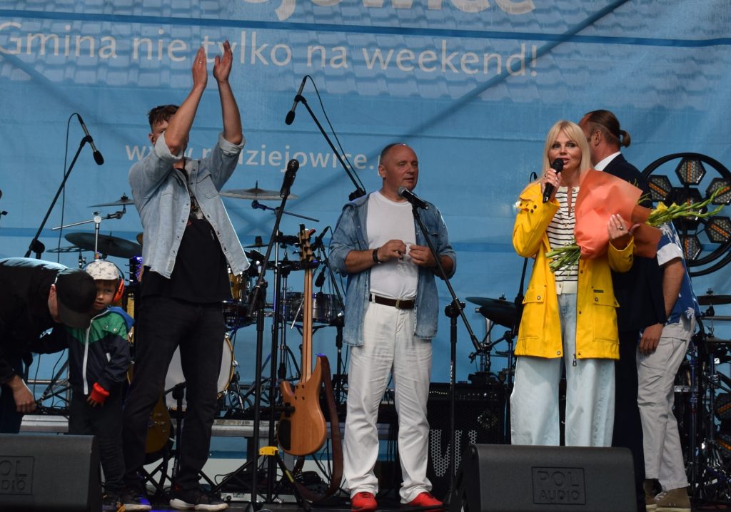 Na scenie podczas koncertu trzy osoby z mikrofonami, w tym kobieta w żółtej kurtce trzymająca bukiet kwiatów. W tle widoczne instrumenty muzyczne i oświetlenie sceniczne.