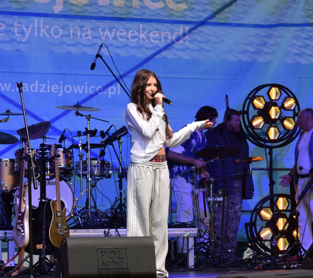 Młoda wokalistka śpiewa na scenie, trzymając mikrofon. W tle widoczni muzycy, instrumenty oraz kolorowe oświetlenie sceniczne.