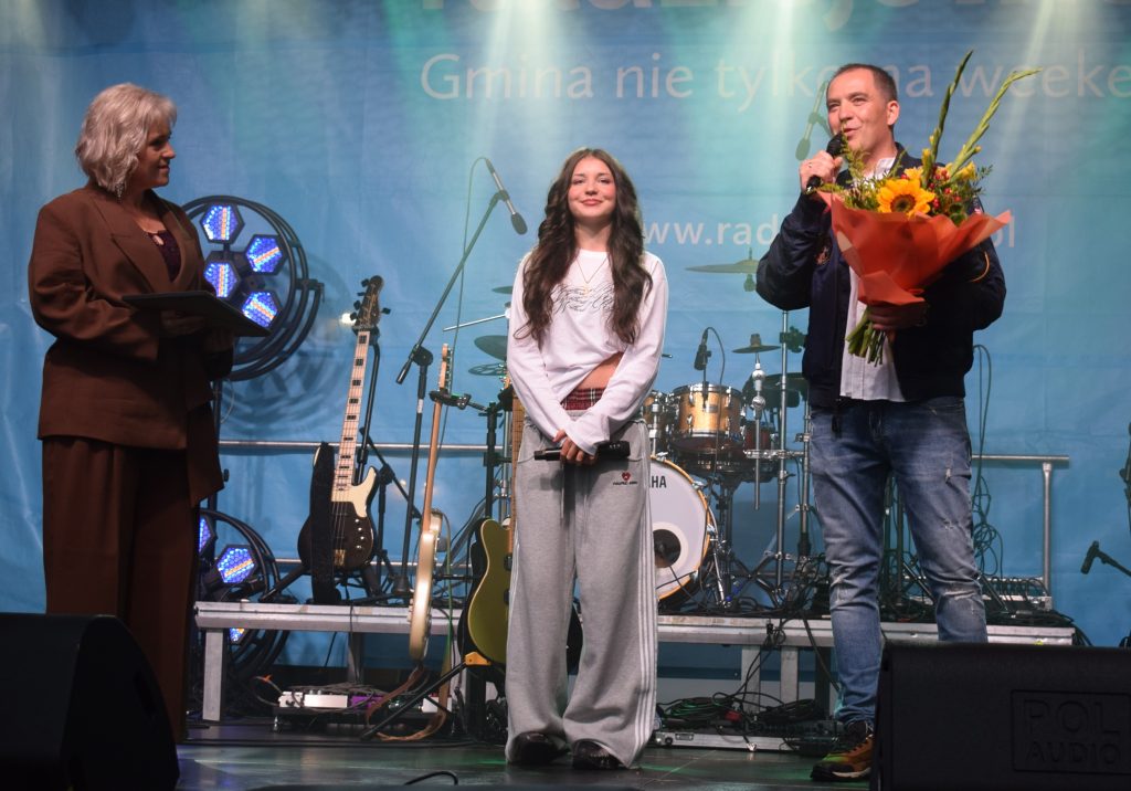Młoda wokalistka stoi na scenie z mikrofonem. Obok niej mężczyzna trzyma bukiet kwiatów, a po drugiej stronie kobieta trzyma dokument. W tle widoczne instrumenty muzyczne.
