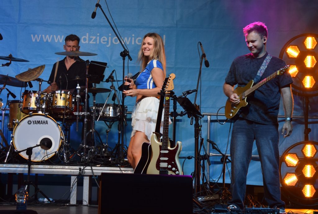 Wokalistka występuje na scenie z zespołem muzycznym. W tle widoczny perkusista i gitarzysta oraz kolorowe oświetlenie sceniczne.