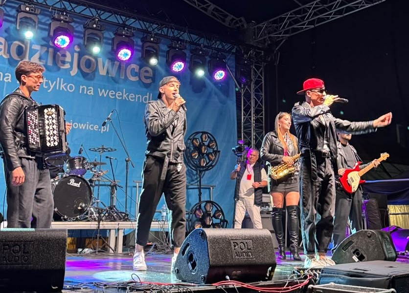 Zespół muzyczny występuje na scenie. Wokalista śpiewa do mikrofonu, obok niego występują muzycy z akordeonem, gitarą i saksofonem.