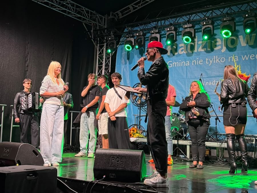 Artysta występuje na scenie z mikrofonem, a obok niego stoją uczestnicy wydarzenia i muzycy. Scenę oświetla zielone światło.