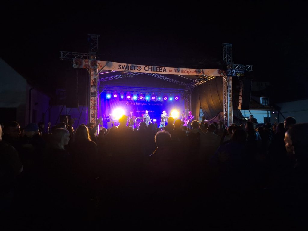 Wieczorny koncert plenerowy. Na oświetlonej scenie występują artyści, a przed sceną zgromadziła się licznie publiczność. Na scenie widoczny jest napis „Święto Chleba w Radziejowicach”.