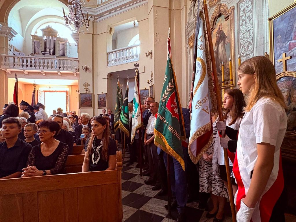 Wnętrze kościoła podczas uroczystości pogrzebowych. Po prawej stronie stoją poczty sztandarowe, a w ławkach siedzą uczestnicy ceremonii.