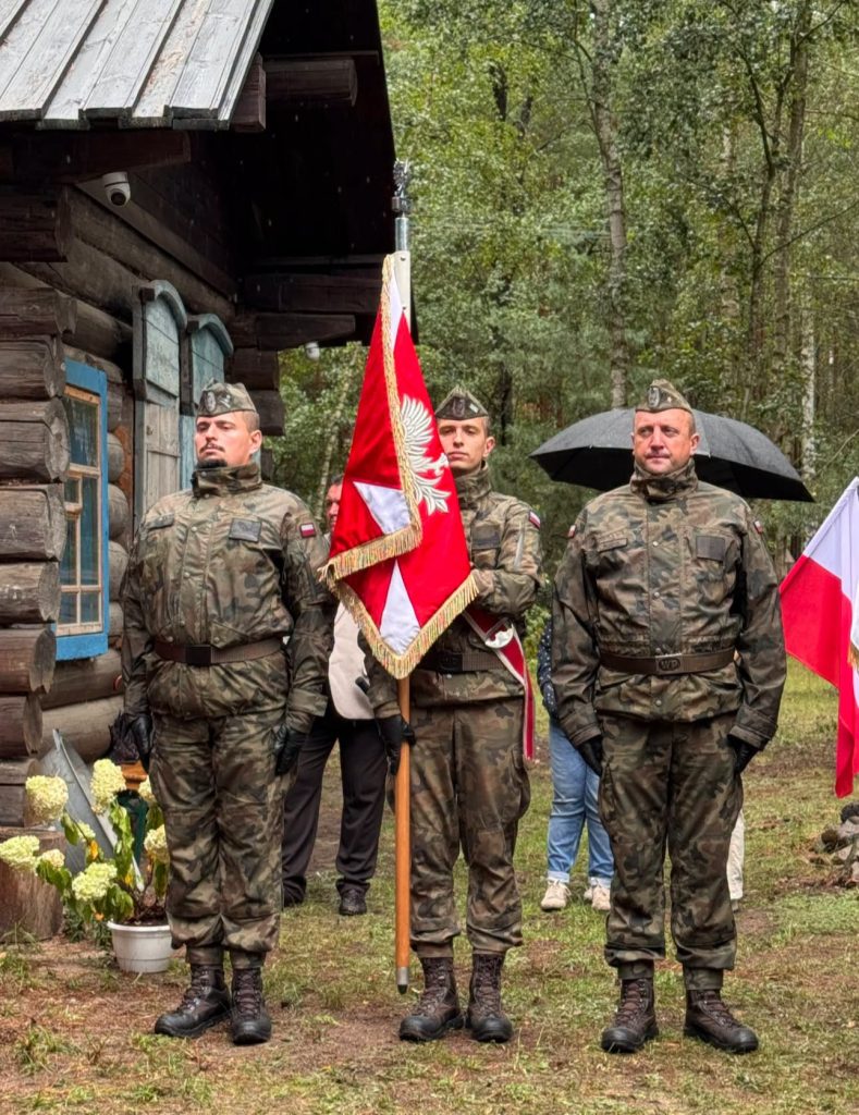 Trzech żołnierzy w mundurach wojskowych stoi przed drewnianą kaplicą. Środkowy żołnierz trzyma biało-czerwony sztandar.
