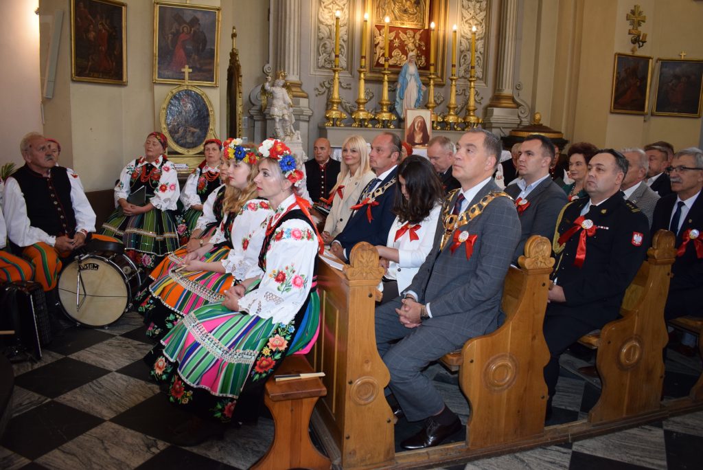 Osoby siedzą na ławkach w kościele podczas Mszy. Na pierwszej ławce i w tle widać osoby ubrane w stroje łowickie