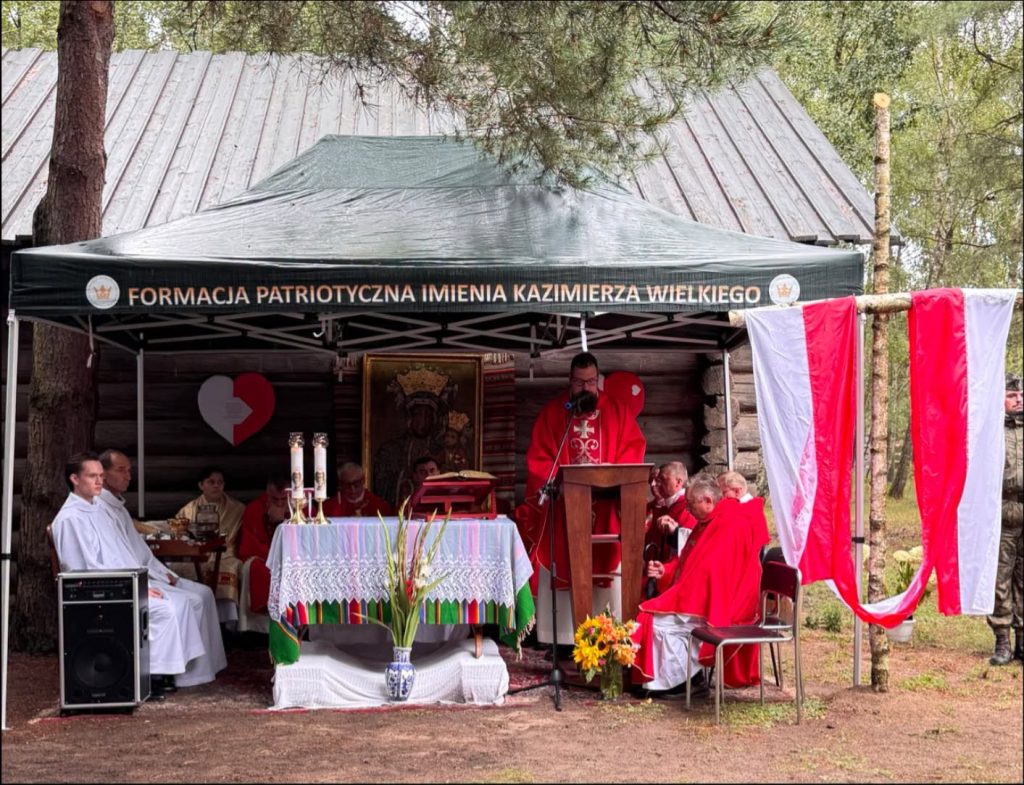 Widok ołtarza polowego podczas uroczystości patriotyczno-religijnej przy drewnianej kaplicy. Przy ołtarzu zgromadzeni są duchowni, a obok powiewają biało-czerwone flagi.