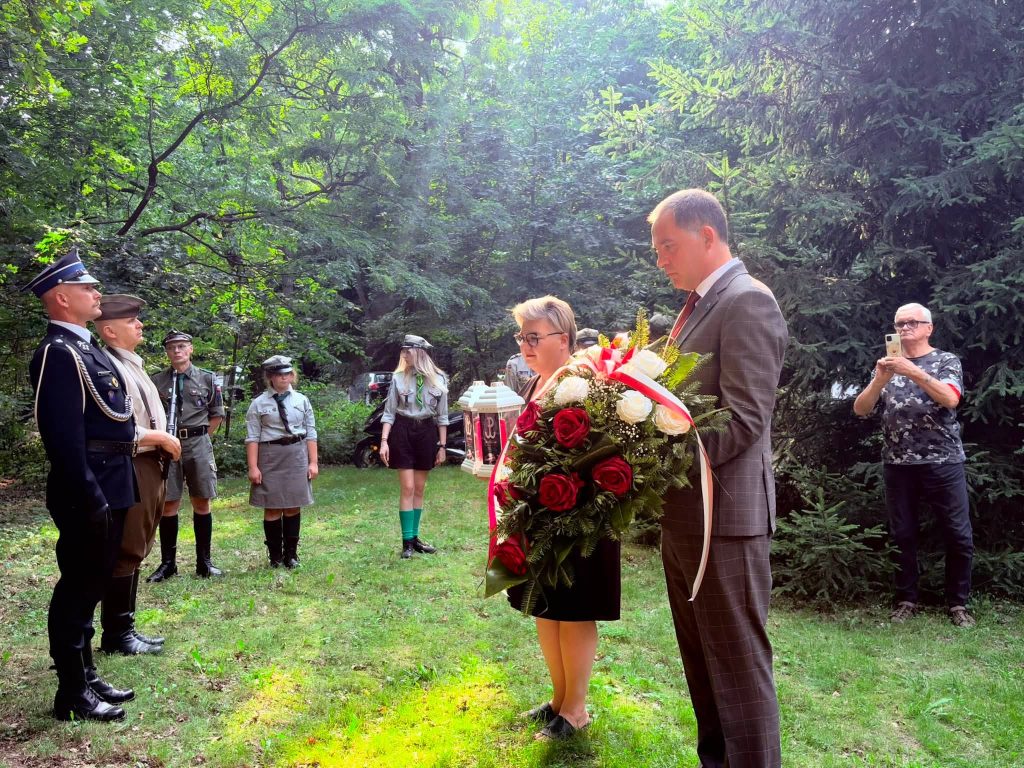 Przedstawiciele samorządu składają wiązankę kwiatów i zapalony znicz przed pomnikiem żołnierzy Armii Krajowej. W tle stoją harcerze i uczestnicy uroczystości.