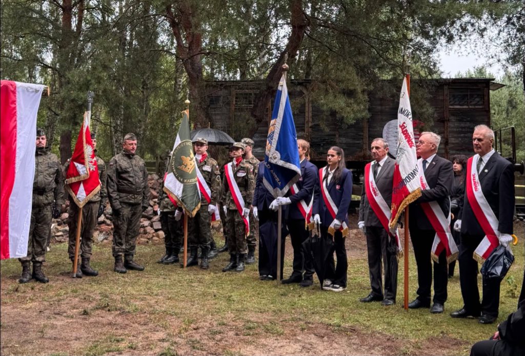 Poczet sztandarowy i uczestnicy uroczystości patriotycznej stoją w lesie przed drewnianą kaplicą. Widoczne są polskie flagi oraz sztandary organizacji i formacji patriotycznych.