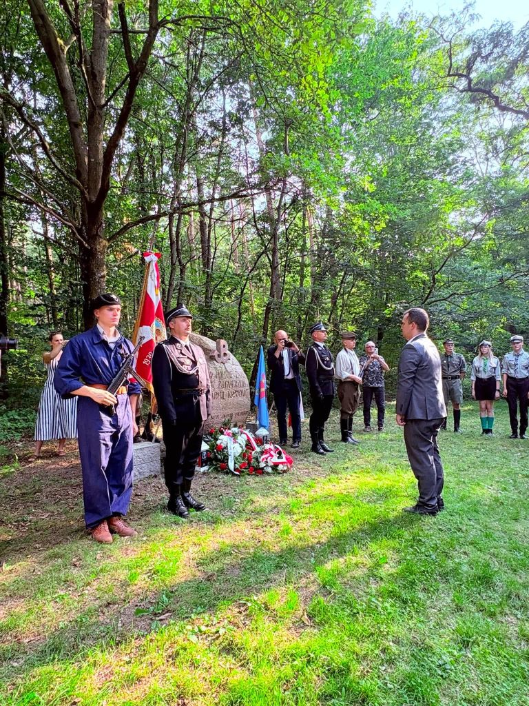 Wójt Gminy Radziejowice oddaje hołd przed pomnikiem żołnierzy Armii Krajowej. Obok stoją członkowie honorowej warty, harcerze i uczestnicy wydarzenia.