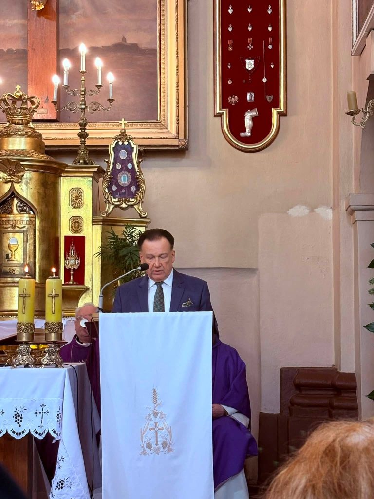 Przy ambonie w kościele przemawia przedstawiciel samorządu. Za nim znajdują się duchowni uczestniczący w mszy pogrzebowej.