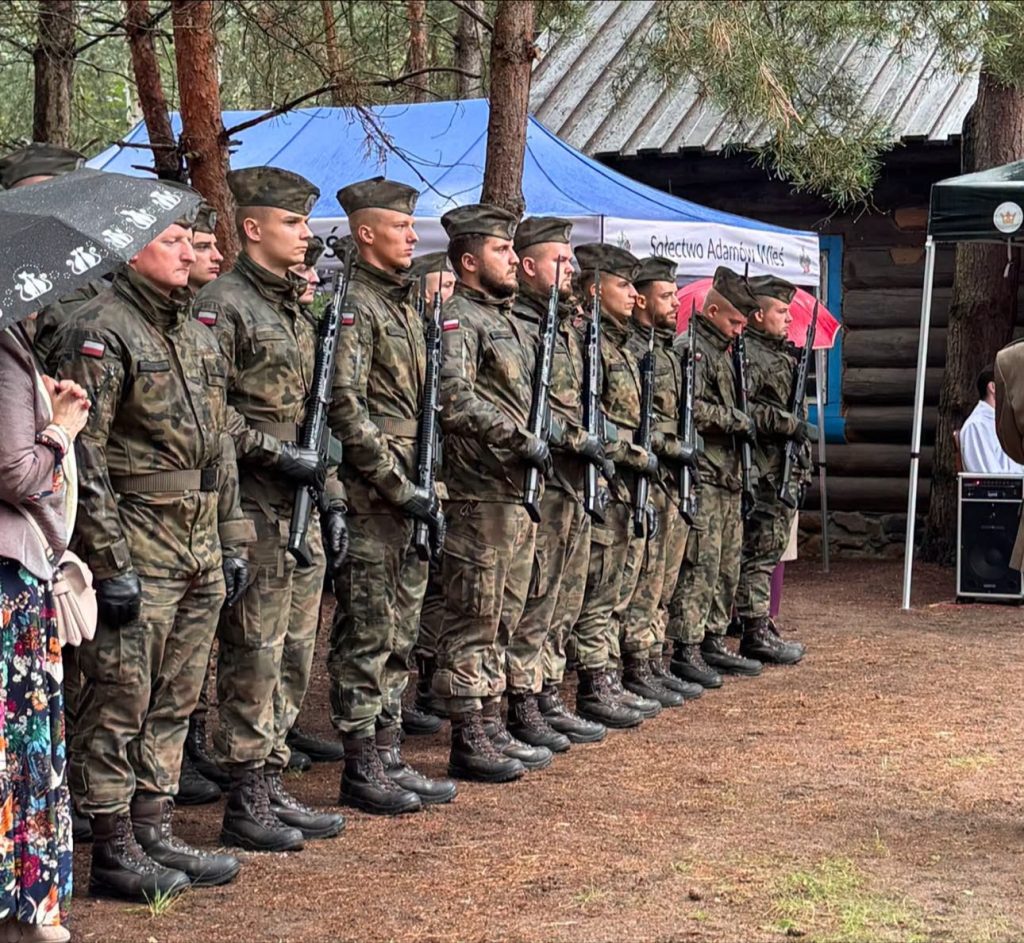 Oddział żołnierzy w mundurach stoi w szeregu podczas uroczystości patriotycznej. Żołnierze trzymają broń, a w tle widoczny jest namiot uczestników wydarzenia.
