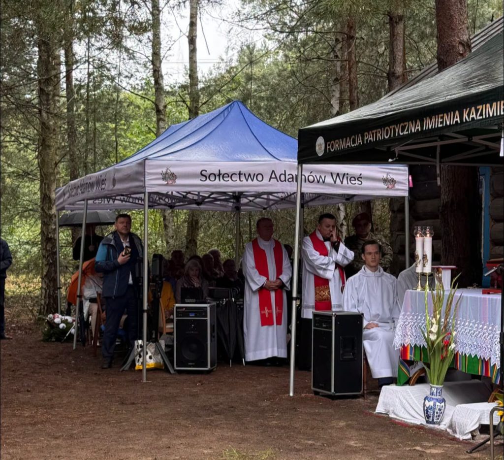 Widok miejsca uroczystości w lesie. Przy ołtarzu polowym znajdują się duchowni, a obok ustawiono namioty dla uczestników wydarzenia.