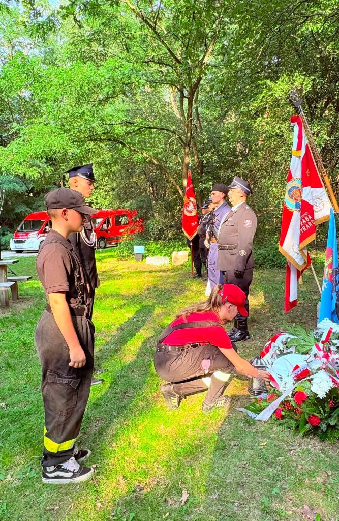 Młoda druhna Ochotniczej Straży Pożarnej zapala znicz przy pomniku upamiętniającym żołnierzy Armii Krajowej. Obok stoją strażacy i członkowie honorowej warty.