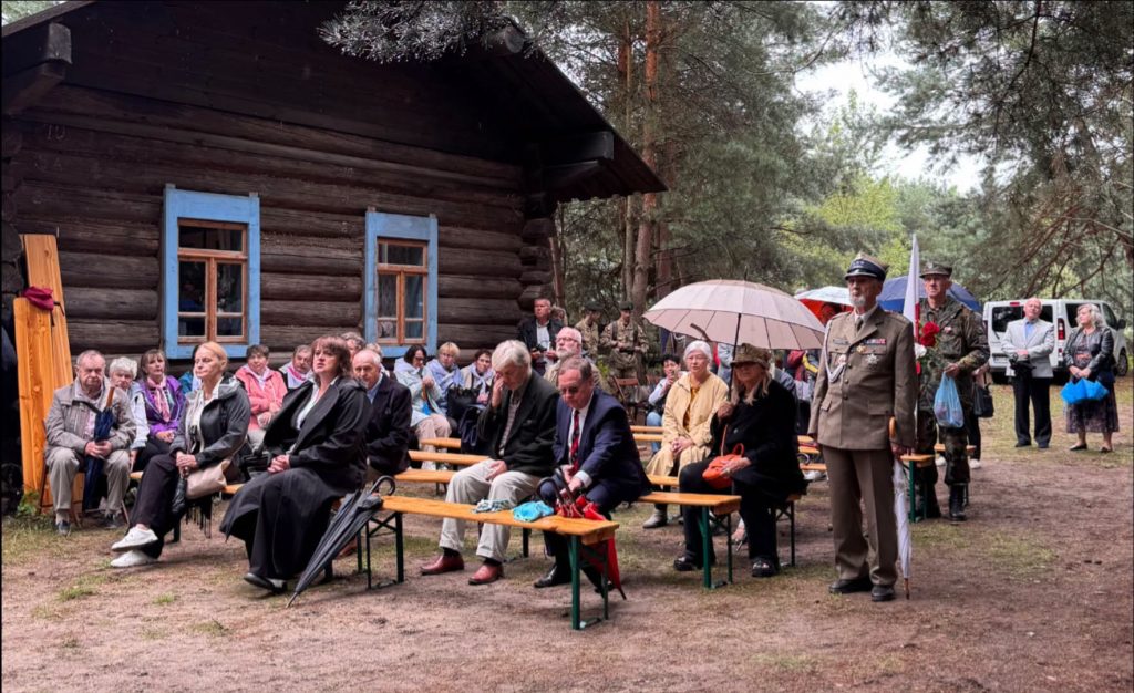 Uczestnicy uroczystości siedzą na drewnianych ławkach przed leśną kaplicą. Wśród zgromadzonych widoczny jest umundurowany przedstawiciel formacji wojskowej.