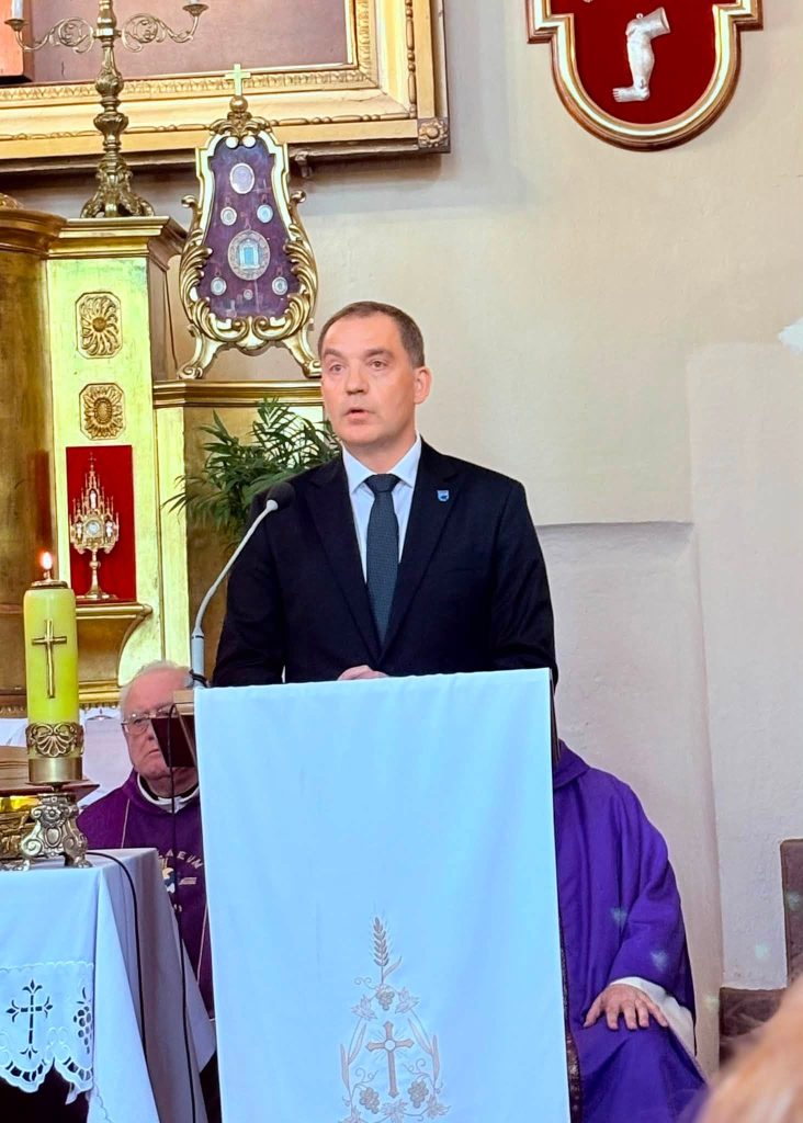 Przy ambonie przemawia Wójt Gminy Radziejowice Artur Jankowski. W tle widoczni są duchowni oraz elementy wyposażenia prezbiterium.