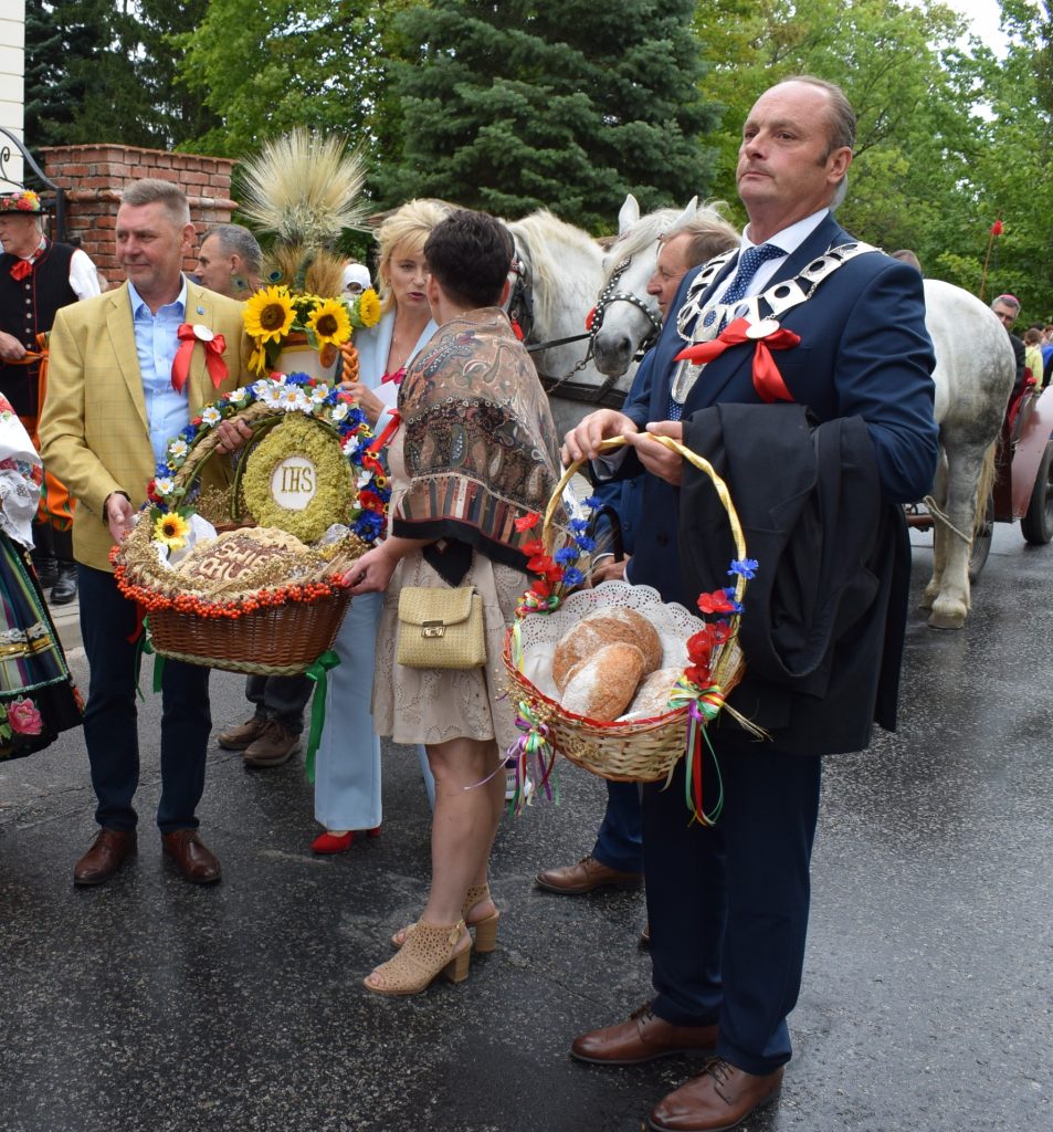 Uczestnicy Święta Chleba prezentują bochenki chleba i ozdobne kompozycje dożynkowe. W tle widoczny jest zaprzęg konny oraz uczestnicy uroczystości.