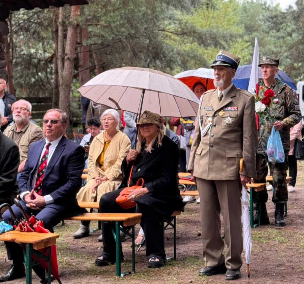 Uczestnicy uroczystości obserwują jej przebieg. Na pierwszym planie stoją osoby siedzące na ławkach oraz umundurowany przedstawiciel formacji patriotycznej.