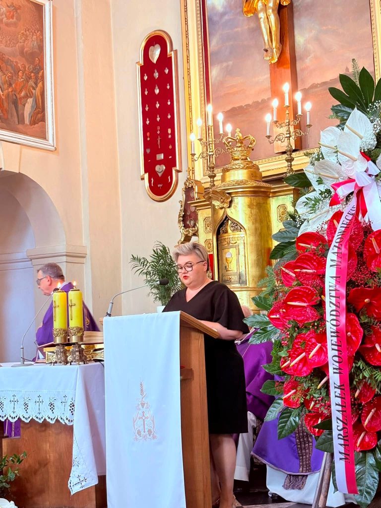 Przy ambonie przemawia Przewodnicząca Rady Gminy Radziejowice Paulina Partyka. Obok znajduje się okazały wieniec pogrzebowy, a w tle widoczni są duchowni.