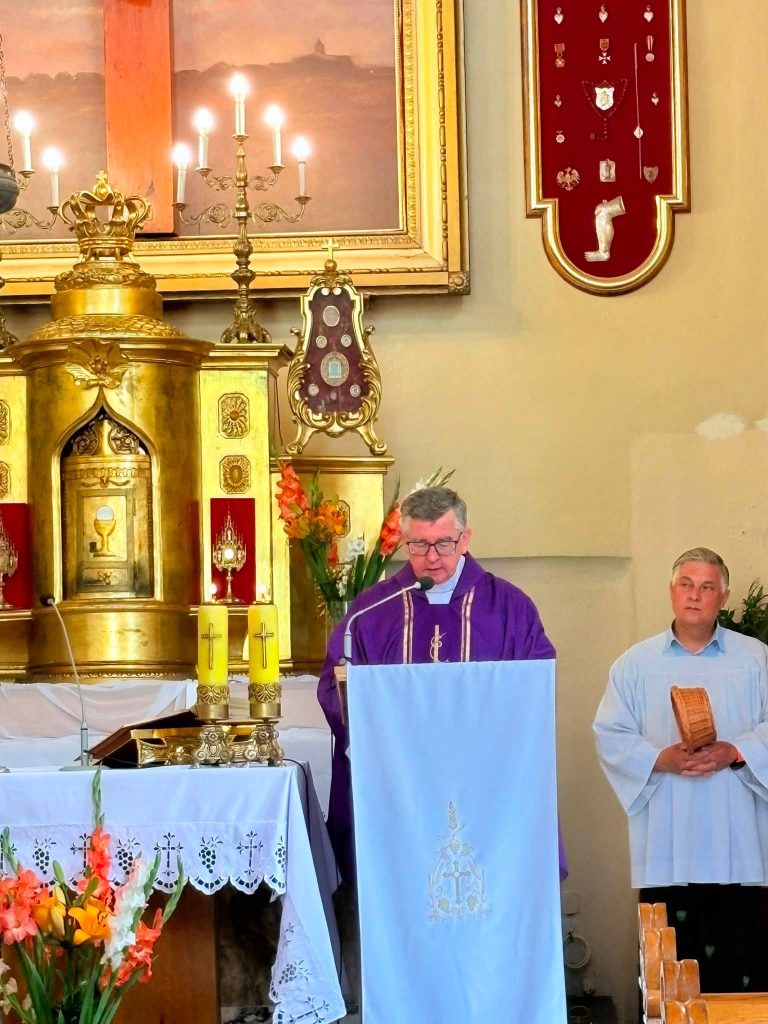 Duchowny prowadzi uroczystość pogrzebową przy ambonie w kościele. Obok niego stoi osoba w stroju liturgicznym, a w tle widoczny jest ozdobny ołtarz i zapalone świece.