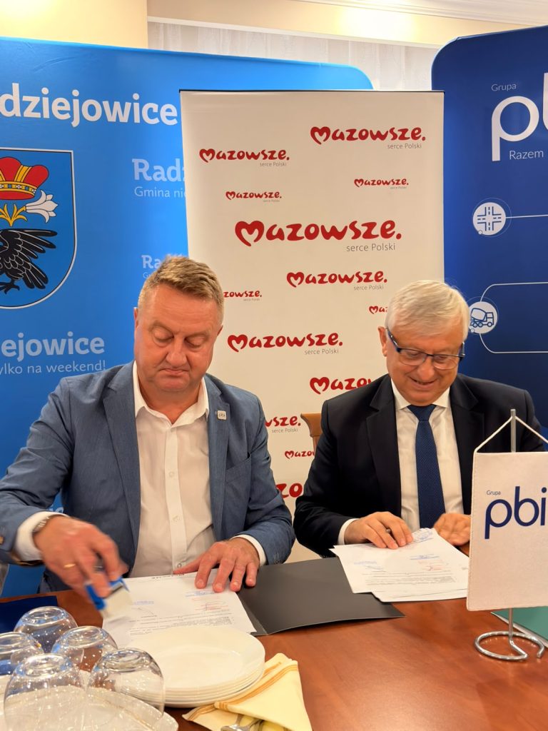 Dwóch uczestników spotkania podpisuje dokumenty przy stole konferencyjnym. W tle widoczne są ścianki promocyjne Mazowsza, Gminy Radziejowice oraz Grupy PBI.