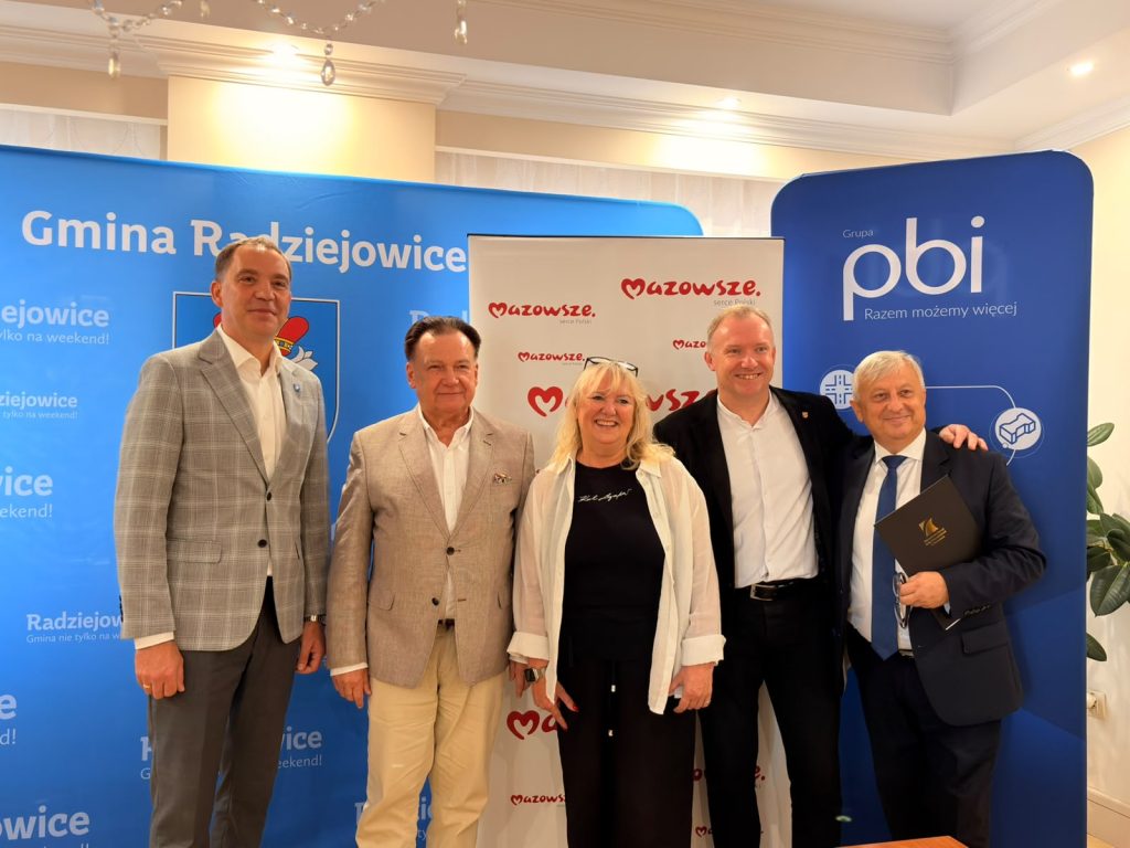 Grupa uczestników spotkania pozuje do pamiątkowego zdjęcia na tle ścianek promocyjnych Gminy Radziejowice, Mazowsza oraz Grupy PBI.