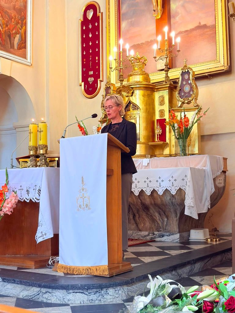 Kobieta przemawia przy ambonie podczas uroczystości pogrzebowej. W tle widoczny jest bogato zdobiony ołtarz, a w dolnej części zdjęcia znajdują się kwiaty.