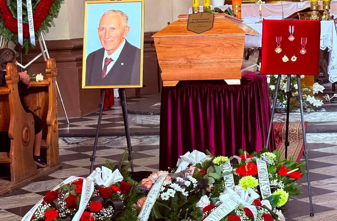 Wnętrze kościoła. Na środku znajduje się trumna śp. Kazimierza Drążkiewicza ustawiona przed ołtarzem. Obok stoi portret zmarłego na sztaludze oraz tablica z odznaczeniami. Przed trumną ułożone są liczne wieńce i wiązanki kwiatów.