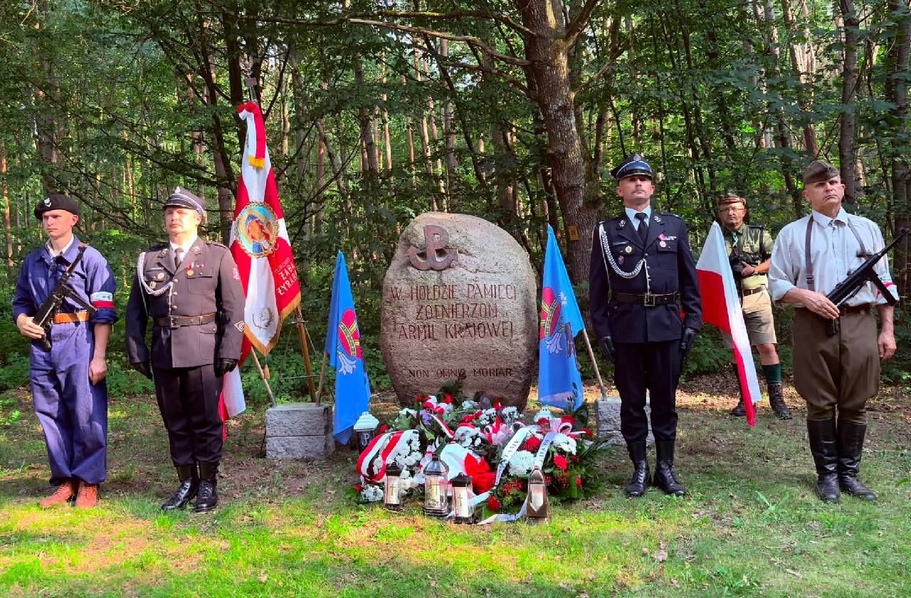 Przed pomnikiem poświęconym żołnierzom Armii Krajowej w lesie stoi honorowa warta w historycznych i strażackich mundurach. U stóp pomnika znajdują się biało-czerwone wiązanki, znicze oraz flagi Polski i Gminy Radziejowice.