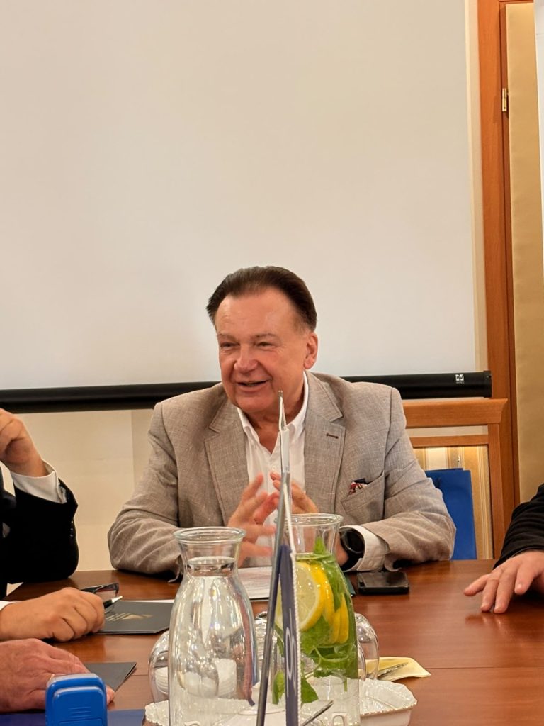 Przy stole konferencyjnym siedzi mężczyzna w jasnej marynarce, przemawiający do uczestników spotkania. Na stole znajdują się karafki z wodą, szklanki oraz dzbanek z wodą i plasterkami cytryny.