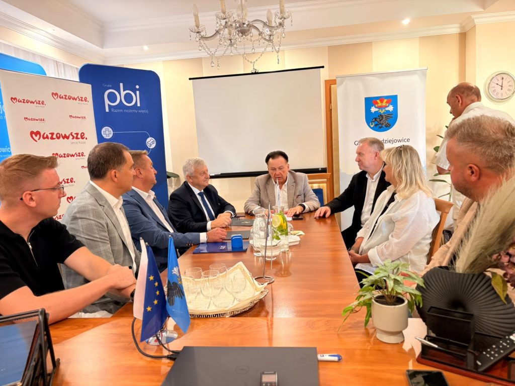 Uczestnicy spotkania prowadzą rozmowy przy stole konferencyjnym. W sali widoczne są materiały promocyjne Gminy Radziejowice oraz partnerów wydarzenia.