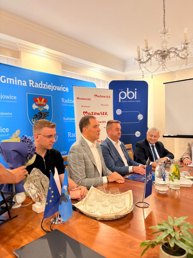 Uczestnicy spotkania siedzą przy stole konferencyjnym. W tle widoczne są ścianki promocyjne Gminy Radziejowice, Mazowsza oraz Grupy PBI.
