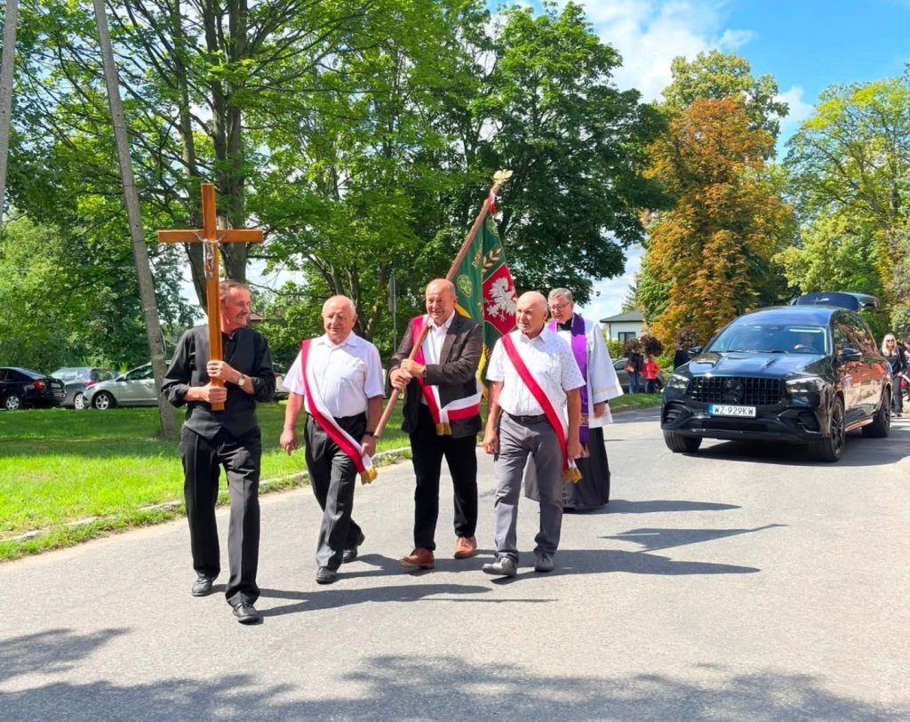 Uczestnicy uroczystości pogrzebowej przechodzą drogą w kondukcie żałobnym. Na czele znajduje się osoba niosąca krzyż oraz osoby ze sztandarem, za nimi idą pozostali uczestnicy i duchowny.