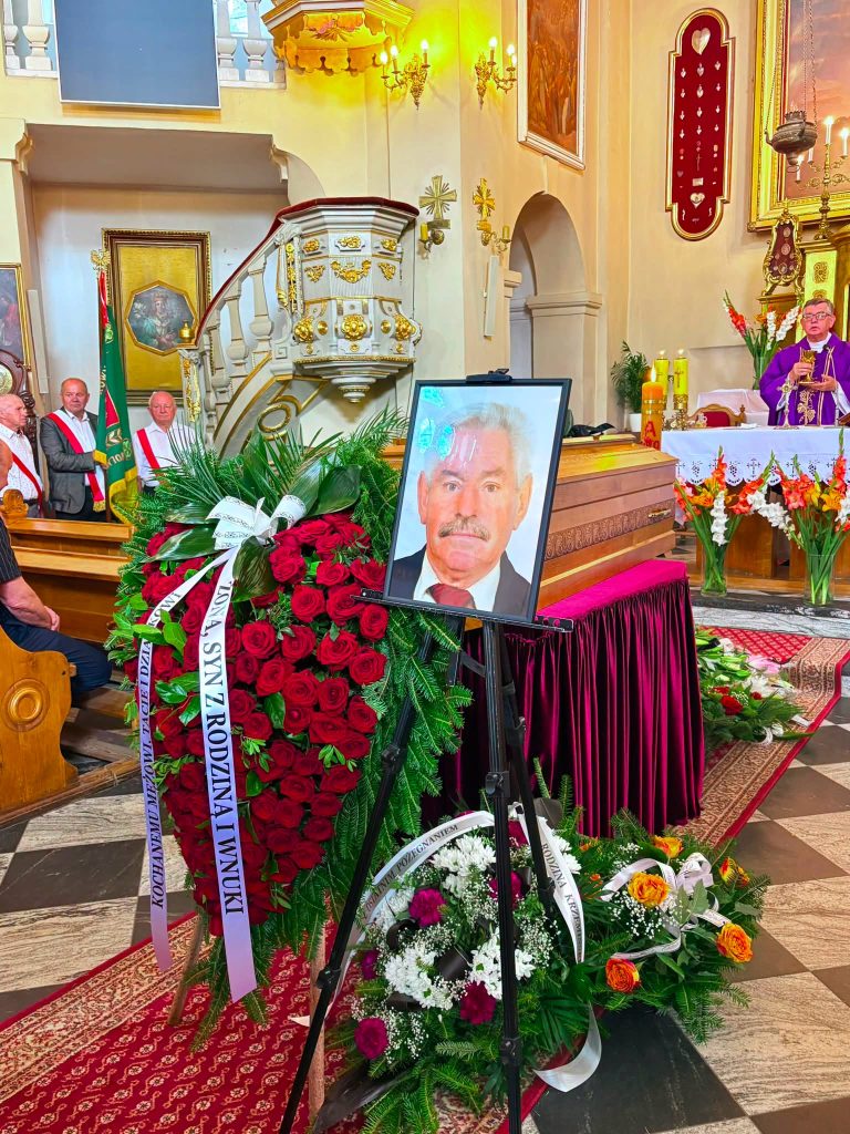 Wnętrze kościoła podczas ceremonii pogrzebowej. Przed ołtarzem znajduje się trumna, portret zmarłego oraz liczne wieńce i kwiaty. W tle widoczny jest duchowny odprawiający uroczystość.
