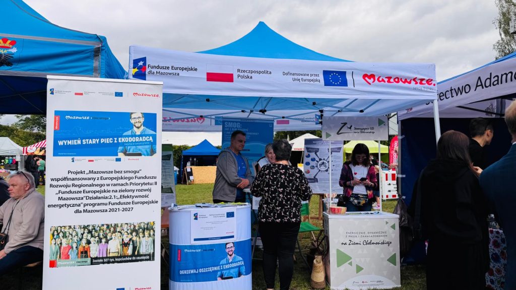 Stoisko projektu „Mazowsze bez smogu” podczas wydarzenia plenerowego. W centrum znajduje się namiot z oznaczeniami Funduszy Europejskich dla Mazowsza i kampanii „Mazowsze bez smogu”, a przed nim uczestnicy rozmawiają z przedstawicielami stoiska i zapoznają się z materiałami edukacyjnymi.