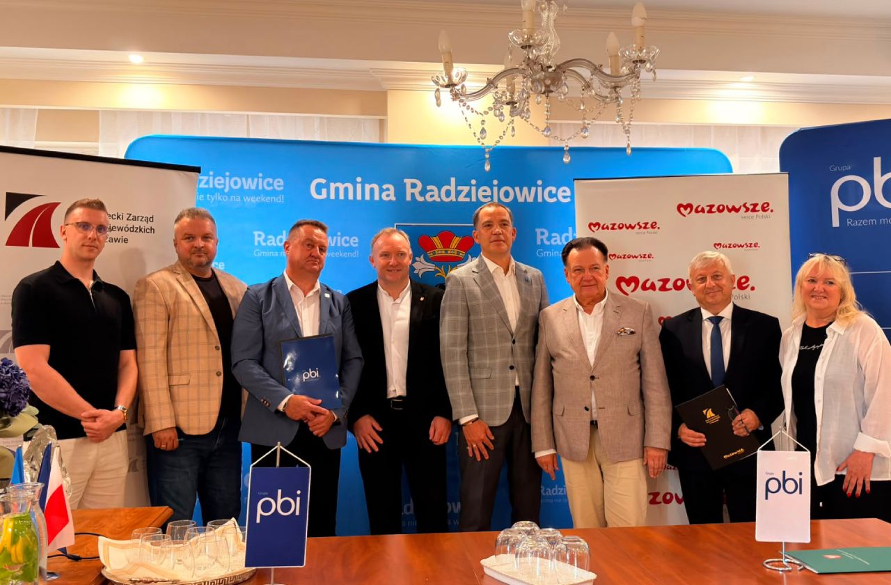 Uczestnicy spotkania pozują do wspólnego zdjęcia po zakończeniu podpisania dokumentów. W tle znajdują się ścianki promocyjne Gminy Radziejowice, Mazowsza i Grupy PBI.