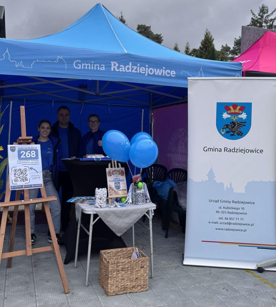 Stoisko promocyjne Gminy Radziejowice pod niebieskim namiotem podczas wydarzenia plenerowego. Przy stoisku znajdują się materiały informacyjne, balony oraz roll-up z herbem gminy.