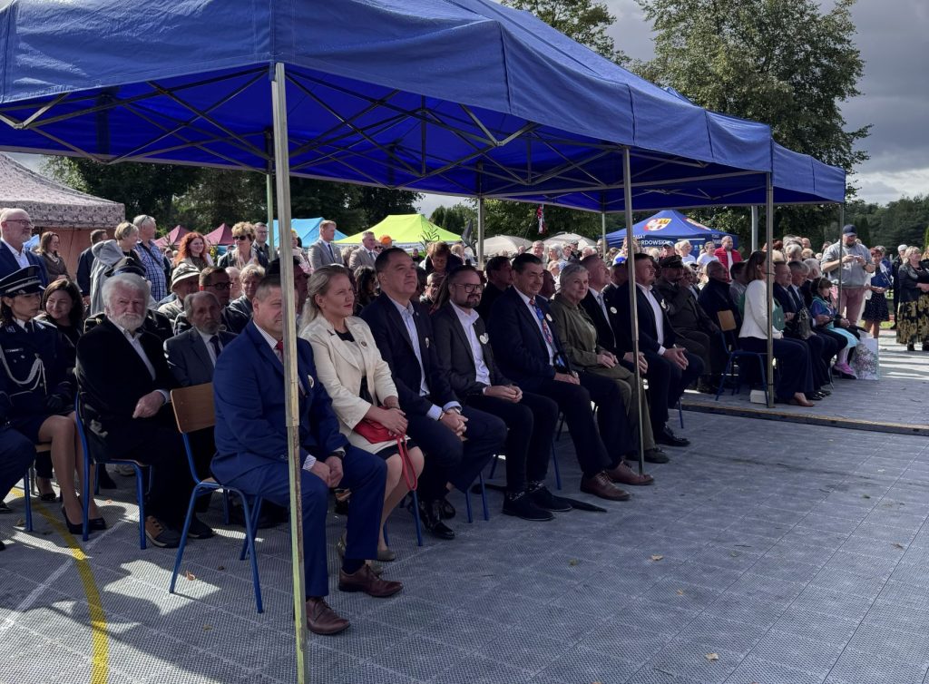 Goście wydarzenia siedzą pod niebieskim namiotem i uczestniczą w uroczystościach dożynkowych.