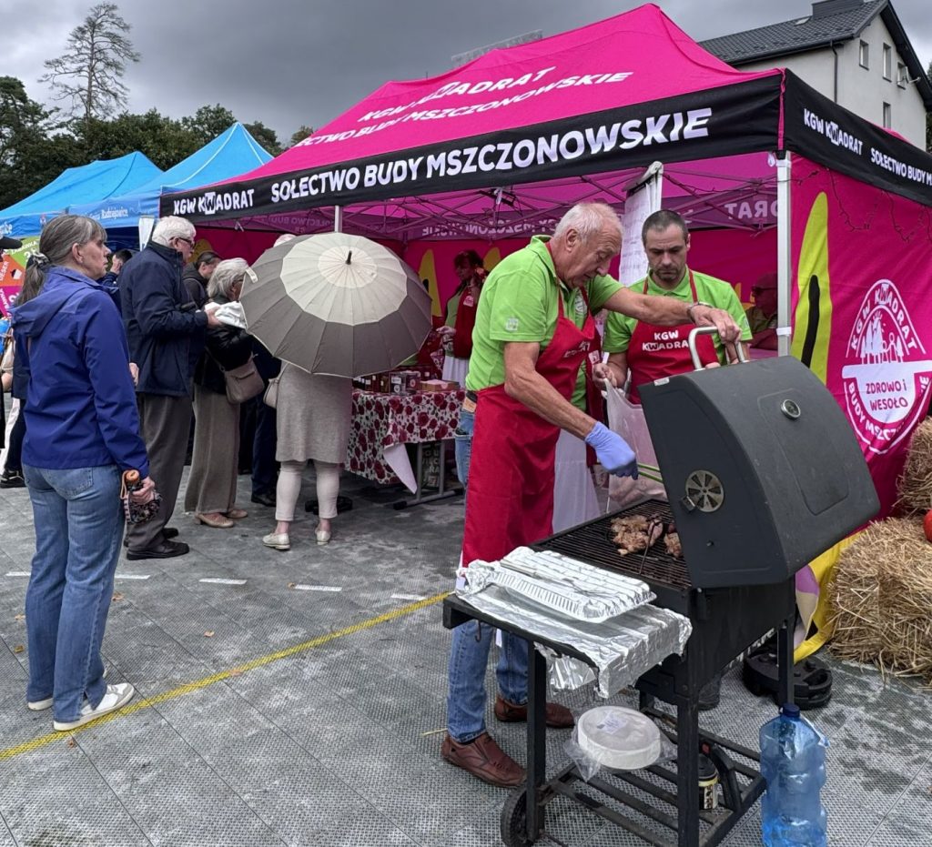 Mężczyźni przygotowują potrawy na grillu przy różowym stoisku Koła Gospodyń Wiejskich podczas wydarzenia plenerowego.
