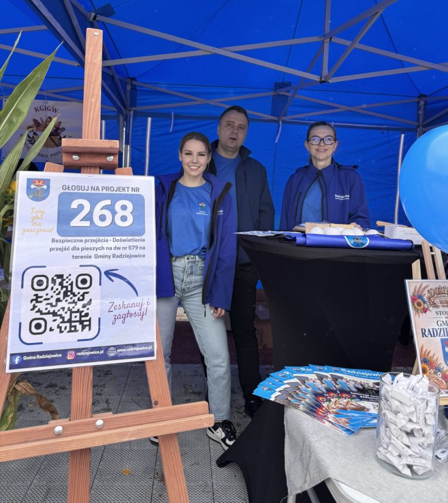 Trzy osoby reprezentujące Gminę Radziejowice stoją przy stoisku promocyjnym z plakatem zachęcającym do głosowania na projekt nr 268 dotyczący bezpiecznych przejść dla pieszych.