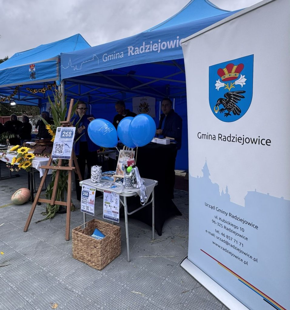 Stoisko promocyjne Gminy Radziejowice podczas wydarzenia plenerowego, z materiałami informacyjnymi, niebieskimi balonami i roll-upem z herbem gminy.Stoisko promocyjne Gminy Radziejowice podczas wydarzenia plenerowego, z materiałami informacyjnymi, niebieskimi balonami i roll-upem z herbem gminy.