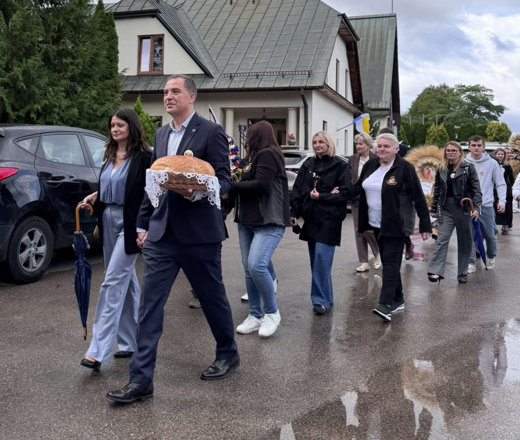 Uczestnicy korowodu dożynkowego idą ulicą w deszczową pogodę. Na pierwszym planie mężczyzna niesie tradycyjny bochen chleba.