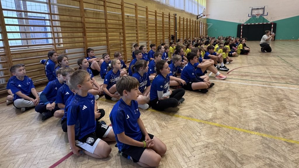 Dzieci w niebieskich i żółtych koszulkach sportowych siedzą na parkiecie sali gimnastycznej i słuchają prowadzącej, która siedzi przed nimi.