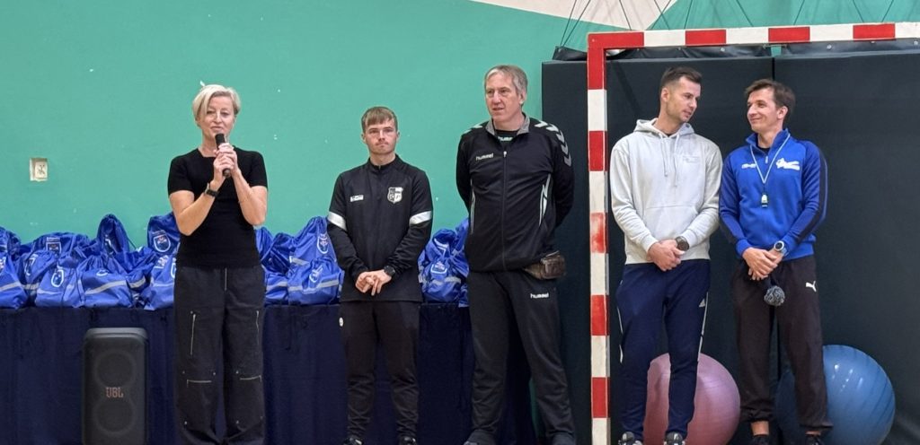 Cztery osoby stoją przed stołem z niebieskimi strojami sportowymi. Kobieta przemawia do mikrofonu, a obok niej stoją trzej mężczyźni w odzieży sportowej.